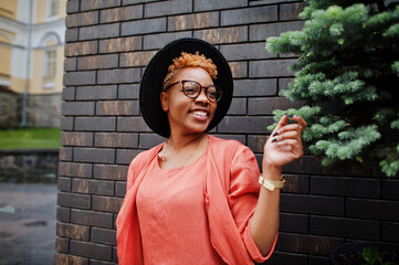 African red haired woman wear eyeglasses, orange dress and black hat posing outdoor.