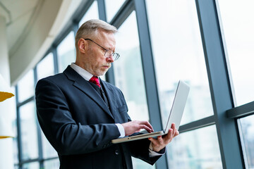 Senior serious businessman standing near window with panoramic city view. Laptop in hands.