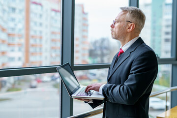 Fototapeta premium Man looking away from window. Laptop in hands, man thinking while checking e-mails standing near window. Photo from the side