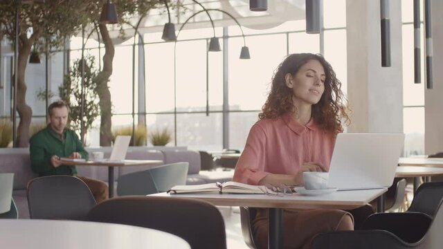 Medium shot of happy Caucasian woman wearing elegant blouse sitting in cafe, working on laptop and enjoying morning coffee while man using PC on background