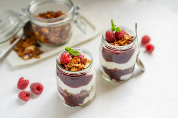 Homemade organic granola or muesli with natural yogurt, fresh raspberries and mint leaf in a jars, white table near window. Healthy beautiful background, sunny light. Selective focus, copy space.