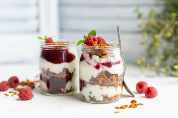 Homemade organic granola or muesli with natural yogurt, fresh raspberries and mint leaf in a jars, white table near window. Healthy beautiful background, sunny light. Selective focus, copy space.