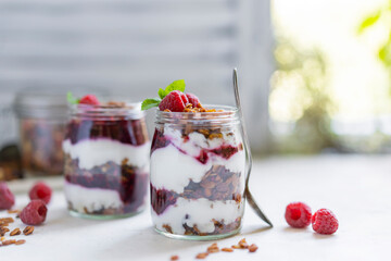 Homemade organic granola or muesli with natural yogurt, fresh raspberries and mint leaf in a jars, white table near window. Healthy beautiful background, sunny light. Selective focus, copy space.