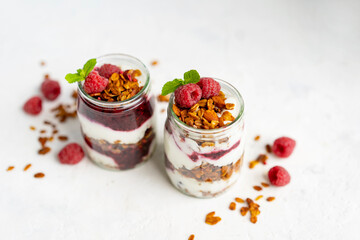 Homemade organic granola or muesli with natural yogurt, fresh raspberries and mint leaf in a jars, white table near window. Healthy beautiful background, sunny light. Selective focus, copy space.
