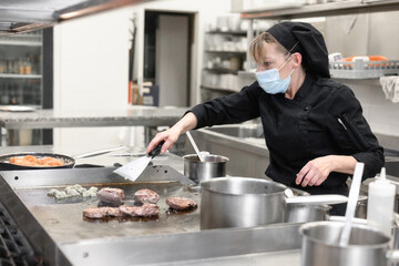 Woman Chef in protective face mask prepare food in the kitchen of a restaurant or hotel. Coronavirus prevention concept. High quality photo