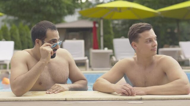 Portrait of young men admiring slim female legs passing by. Handsome Middle Eastern and Caucasian male friends resting at luxurious resort on sunny summer day outdoors at poolside.