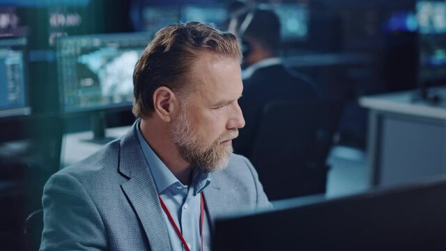 Portrait of Serious Professional Technical Controller Sitting at His Desk with Multiple Computer Displays Before Him. In the Background His Colleagues Working in System Control and Monitoring Center.