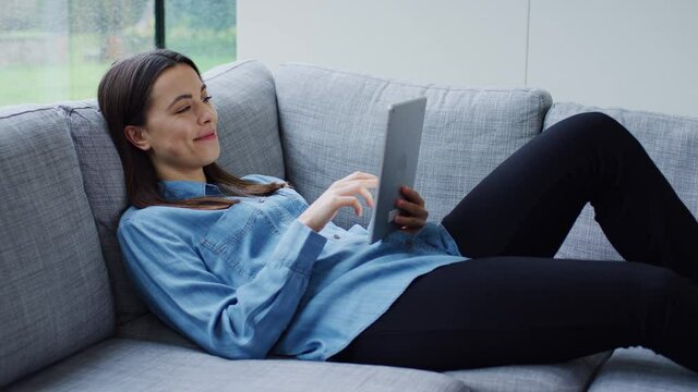 Relaxed young woman at home lying on sofa looking at digital tablet on rainy day and laughing - shot in slow motion