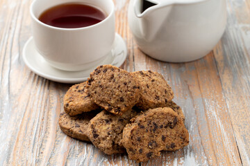 A pile of small american cookies with chocolate chips on a wooden  table, close up, teapot and cup on background
