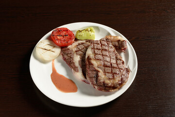 Two fried marble beef steaks with grilled vegetables of tomato, onion and bell pepper on a round plate on a wooden table. Photos for restaurant and cafe menus