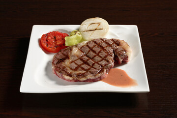 Fried marble beef steaks with grilled vegetables of tomato, onion and bell pepper on a square plate on a wooden table. Photos for restaurant and cafe menus