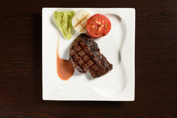 Roasted meat of marble beef steaks with grilled vegetables of tomato, onion and bell pepper on a square plate on a wooden table. Top view. Photos for restaurant and cafe menus