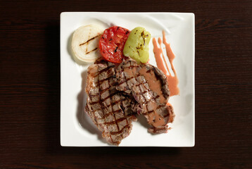 Roasted beef tenderloin with grilled vegetables of tomato, onion and bell pepper on a square plate on a wooden table. Top view. Photos for restaurant and cafe menus