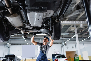 Fototapeta premium low angle view of mechanic in cap and overalls holding wrench while repairing car in service station