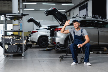 cheerful mechanic in cap waving hand and holding paper cup near cars in workshop