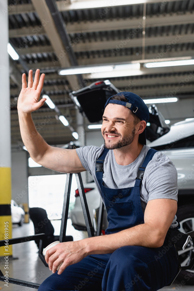 Wall mural cheerful mechanic in cap waving hand and holding paper cup in car workshop