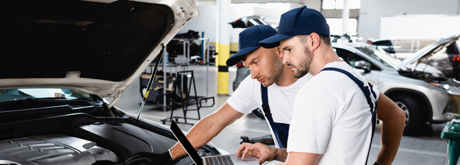 Panoramic shot of auto mechanics looking at laptop screen near car at service station