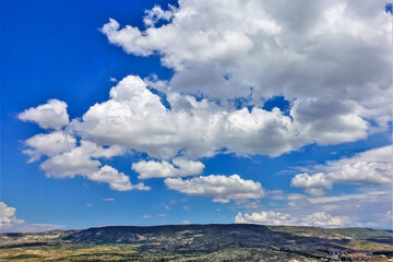 Beautiful white cumulus clouds in the blue sky over Cappadocia. Below is a valley, hills, village. Summer sunny day.