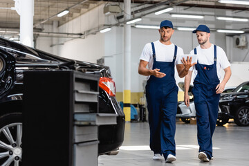 selective focus of handsome mechanics in uniform using digital tablet and walking near cars in workshop