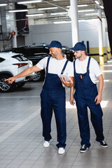 happy mechanic in uniform holding digital tablet and pointing with finger at car near coworker in workshop