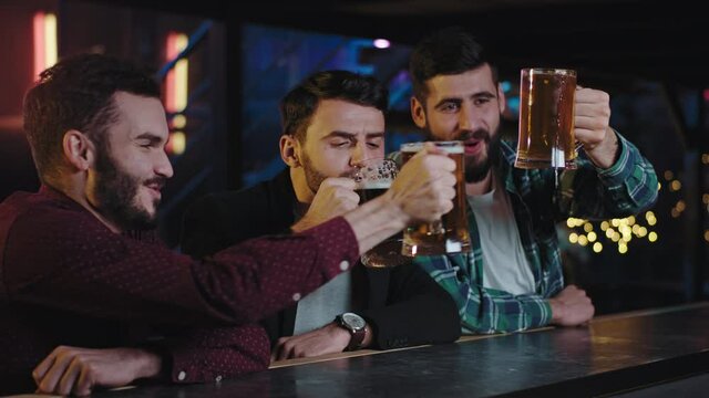 Group of friends charismatic guys in the pub sitting at bar table start drinking big glasses of fresh beer they cheers with glasses and feeling so relaxed