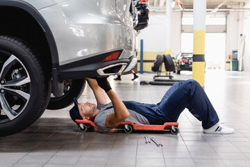 handsome mechanic in uniform and cap lying under car in service center