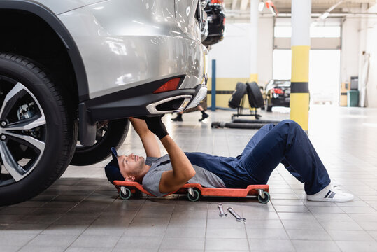 Handsome Mechanic In Uniform And Cap Lying Under Car In Service Center