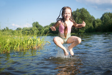 a little girl in a bathing suit jumps and splashes water in the lake in summer