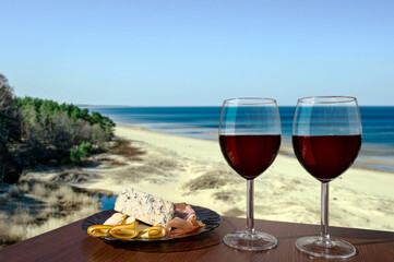 Two glasses of wine with charcuterie assortment with view of beach in Baltic sea. Glass of red wine with different snacks - plate with ham, sliced, blue cheese. Romantic celebration.