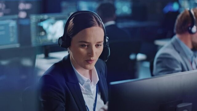 Happy Confident Technical Customer Support Specialist Having a Headset Call while Working on a Computer in a Dark Monitoring and Control Room Filled with Colleagues and Display Screens.