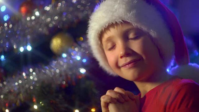 Boy in a Santa hat is dreaming Christmas gifts on  Christmas.  Happy 8 year old child is preparing for the New Year. 4K video of a smiling boy  is praying near Christmas tree about christmas gifts