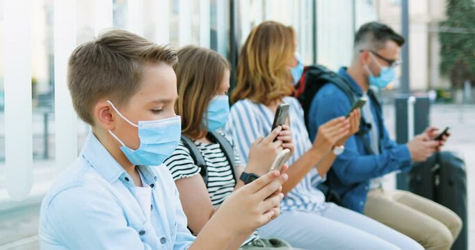 Caucasian Family With Children In Medical Masks Sitting At Bus Stop, Waiting For Transport. Boy And Girl With Mother And Father Using Phones. Kids And Adults Tapping And Scrolling On Smartphones.