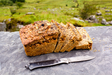 Sliced loaf of the fresh traditional Irish soda bread outside with knife aside and visible greenery on the background. 