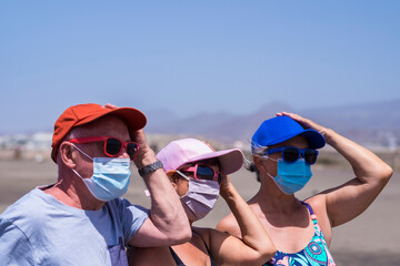 Group of three senior people enjoying the beach together holding one hand on the hat because of the wind, wearing the coronavirus protection mask - active retired seniors and vacation concept