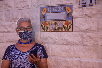 Senior woman standing against  a wall with flower mask because of coronavirus - using smart phone - concept of active elderly during retirement - spanish word for mail boxe