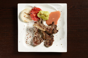 Barbecue beef ribs with grilled vegetables of tomato, onion and bell pepper on a square plate on a wooden table. Top view. Photos for restaurant and cafe menus