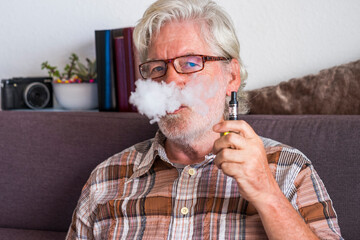 Bearded senior man smoking electronic cigarette sitting on sofa at home - new technology for more...