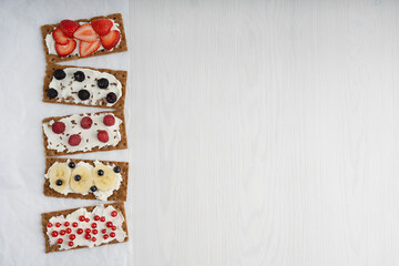 Five pieces of crisp bread with cream cheese and variety of berries such as strawberry, blackberry, raspberry, red currant are laying in a row on white wooden table. Image with copy space