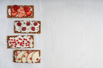 Fototapeta na wymiar Top view of four pieces of vegetarian crisp bread with cream cheese and assortment of red berries such as strawberry, raspberry and red currant laying on white wooden table. Image with copy space