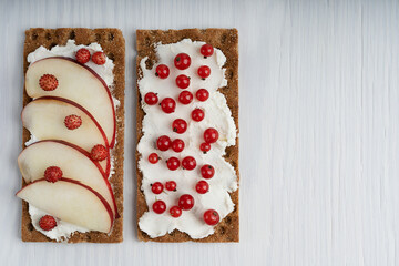 Top view of two crispy bread pieces with cream chese and various berries such as red currant and wild strawberry laying at the left side on white wooden table at kitchen. Image with copy space