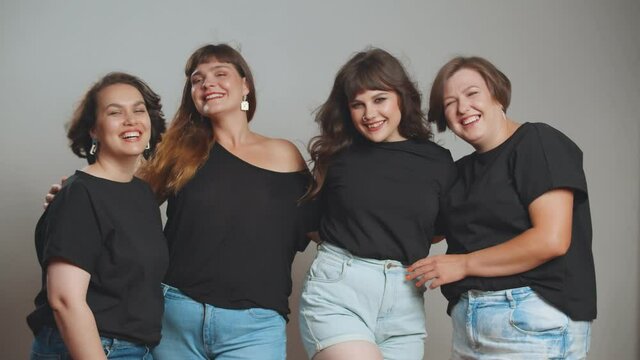 Cheerful plus size women in black t-shirts and jeans embracing and smiling at camera
