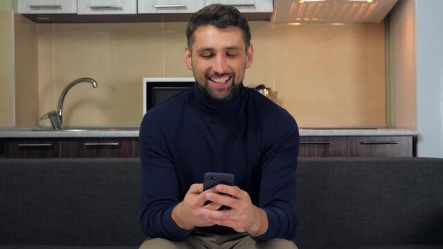 Joyful handsome man holds smartphone in hand communicates in messenger uses smartphone typing message laughs. Guy sits on couch home presses fingers on phone screen writes post social networks, smiles