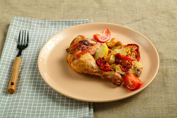 Baked chicken leg with potatoes and tomato on a beige plate on a tablecloth next to a fork and napkin.