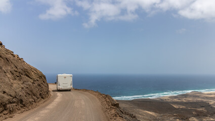 Photo of a motorhome driving along a narrow dirt road in the mountains of the Jandia peninsula. In the background with blue sky and wild cofete beach