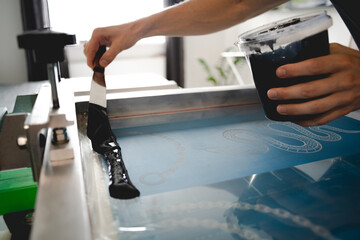Cropped view of artisan applying plastisol ink with spatula on stencil on screen printing machine...