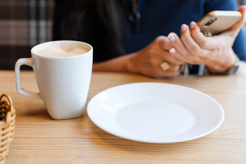 A white coffee Cup and an empty plate on a wooden table against the background girl's hands holding a smartphone while waiting for breakfast in the morning.