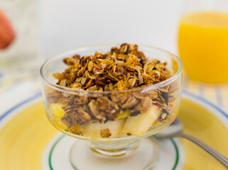 Homemade oat and nut granola on cut up fruit in glass goblet bowl with hint of milk carton and orange juice in the background