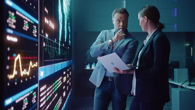 Project Manager and Computer Science Engineer Talk while Using Big Screen Display and a Laptop, Showing Infrastructure Infographics Data. Telecommunications Company System Control and Monitoring Room.