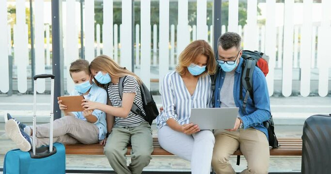 Caucasian family in medical masks sitting on bench with suitcases and waiting for bus at stop outdoor. Kids, brother and sister playing on tablet device. Parents using laptop computer for route plan.