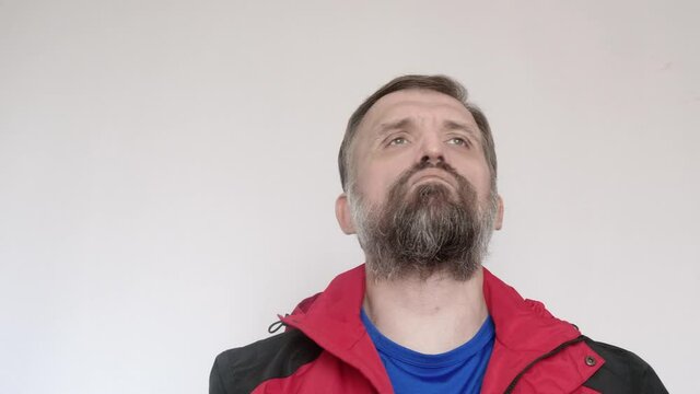 Man with white wall behind him puts on red coat and black baseball cap.